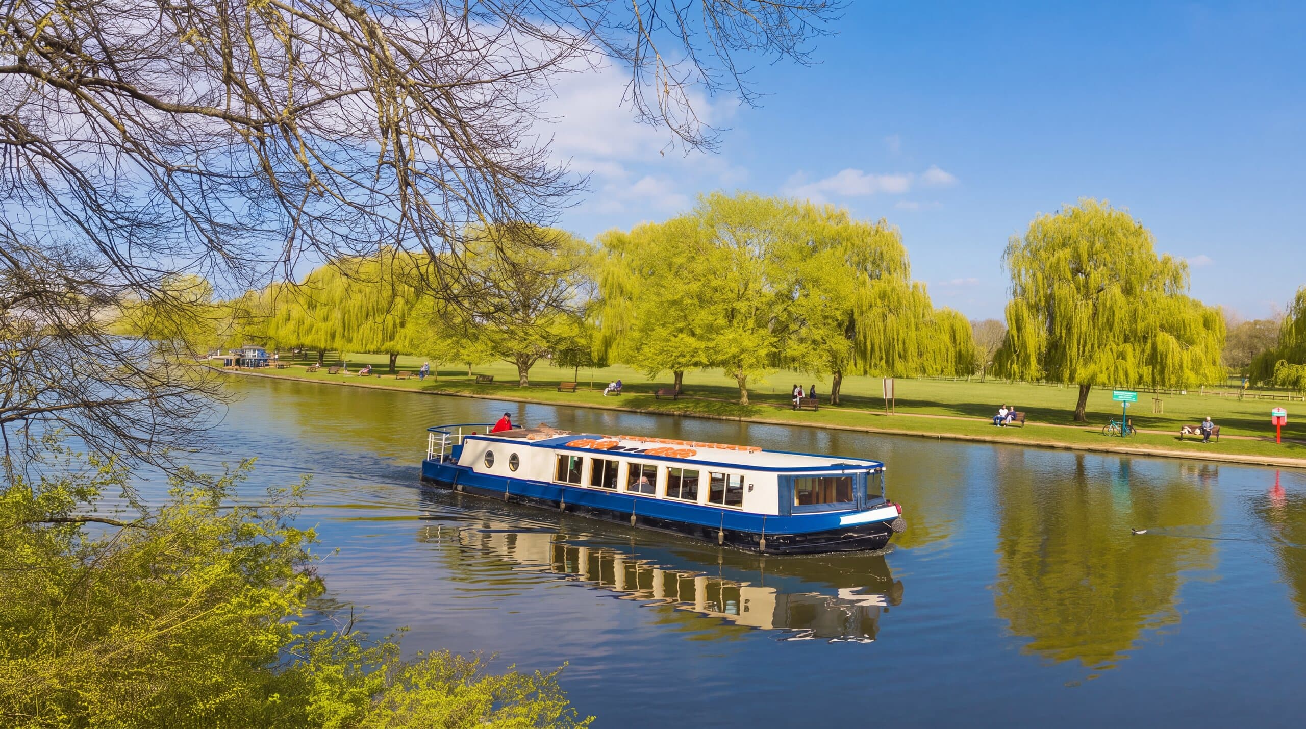 The Rita Ellen, Bancroft Cruisers' blue and white event cruiser, on the River Avon beside willow-lined parkland on a bright day